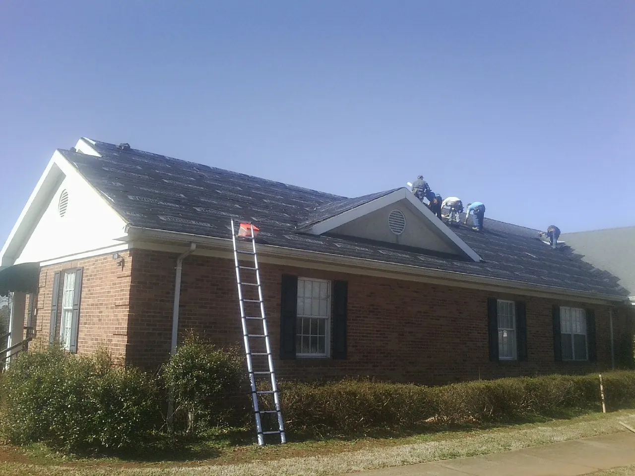 Crew tearing shingles off a brick ranch roof with a ladder set — Emergency Roof Repair work by our crew in Hagaman