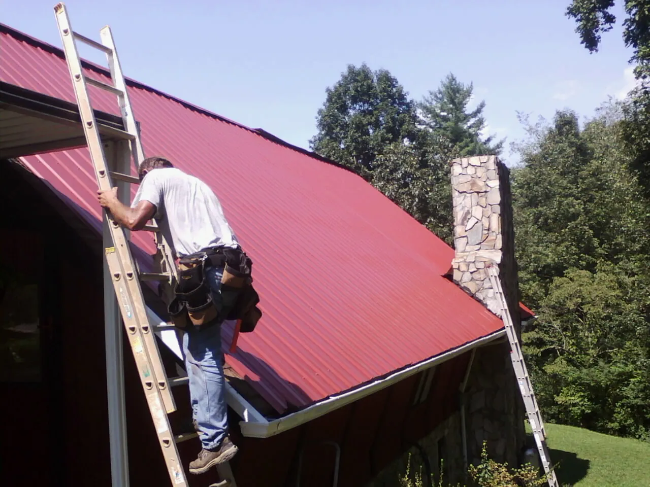 Roofer on a ladder beside a red metal roof and stone chimney — Soffit & Fascia Repair by Roofing Brothers in Hagaman